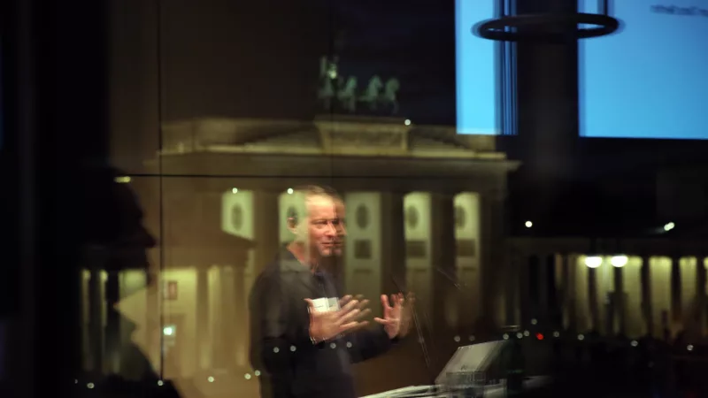 Blick aus dem Fenster auf das Brandenburger Tor, überlagert von einer Reflektion im Fensterglas, die einen Redner am Redner*innenpult spiegelt, der im Innenraum seinen Vortrag hält.