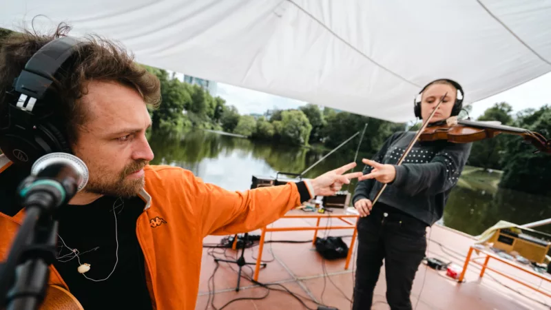 Ein überdachter Steg, an einem Fluss. Darauf zwei Musiker*innen. Eine Person am Mikrofon mit Kopfhörer, eine Person mit Kopfhörern und Geige. Beide sind einander zugewandt und spielen Schere Stein Papier.