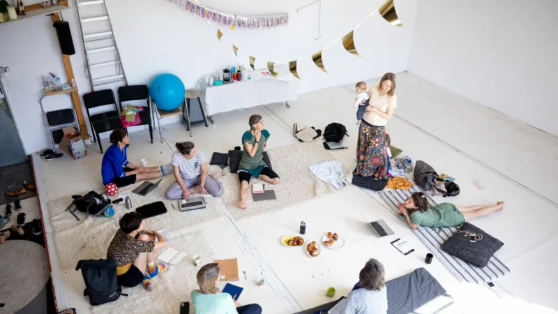 Ein hoher Raum, aus einer Empore herunter fotografiert. Auf dem Boden liegen Decken, im losen Kreis liegen Frauen mit Kindern, Kissen, Laptops. In der Mitte des Kreises stehen Speisen. Eine Frau steht und trägt ihr Kind auf dem Arm.