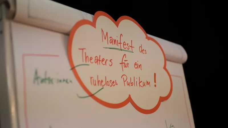 Ein Flipchart auf dem am oberen Ende eine Moderationskarte klebt. Darauf ist zu lesen „Manifest des Theaters für ein ruheloses Publikum“.