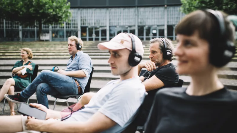 Außenbereich vor einer Halle. 5 Personen sitzen in Vordergrund auf Stühlen, tragen Kopfhörer und schauen sich etwas an, das nicht im Bild ist. Man sieht sie von der Seite.