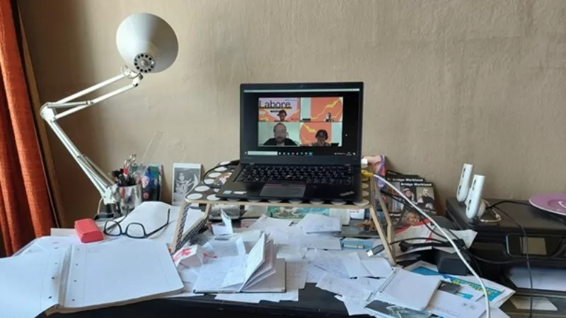 Ein Schreibtisch. Über und über mit Papier, Notizblöcken und ähnlichem Schreizubehör bedeckt. Etwas erhöht steht ein Laptop auf dem eine Online-Konferenz mit drei weiteren Personen stattfindet.