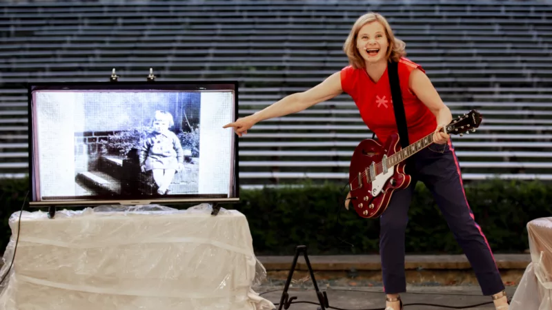 Bernadette La Hengst hat eine E-Gitarre umgehängt und zeigt auf einen Bildschirm, der neben ihr steht. Darauf ist eine schwarz-weiß Fotografe mit einem Kleinkind abgebildet.