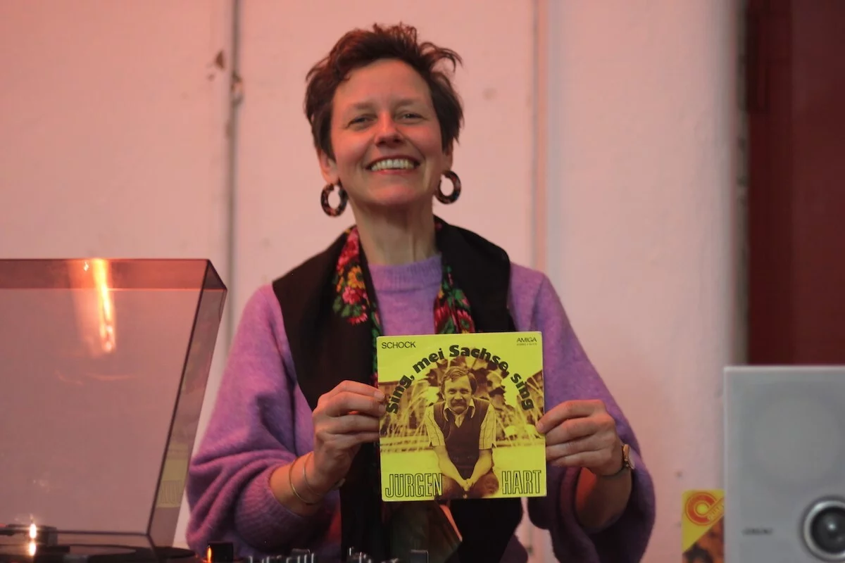 A woman with short hair and striking earrings holds an original Amiga single record in front of her. The title of the record is "Sing mei Sachse sing".