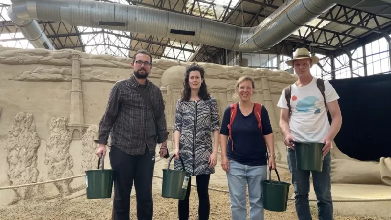 Das Team von Markus&Markus steht, mit Eimern in den Händen, vor großen Sandskulpturen in einer großen Halle in Travemünde.