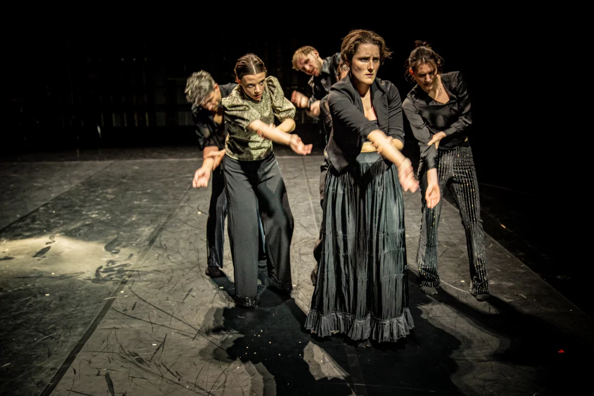 A group of five darkly dressed people stand in the illuminated black stage space. Their posture seems tense, their bodies are sometimes hunched over, each of them supports their right arm stretched out in front with their left hand.