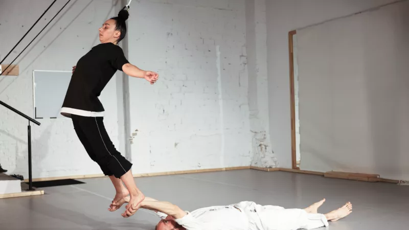 A man dressed in white is lying on his back on the floor of a studio. With outstretched arms, he reaches for the feet of a woman dressed in black, hanging from the ceiling by her hair. Her body is arched over, her arms outstretched.