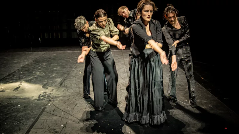 A group of five darkly dressed people stand in the illuminated black stage space. Their posture seems tense, their bodies are sometimes hunched over, each of them supports their right arm stretched out in front with their left hand.