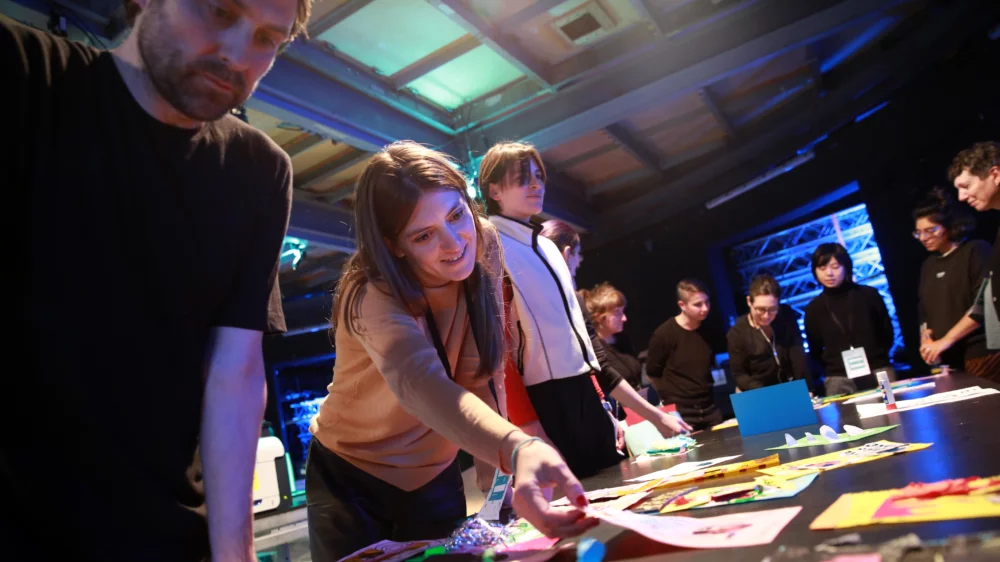 A group of people stand lined up around a large work table with documents and colored pieces of paper at the zine workshop. One of them reaches for a sheet.