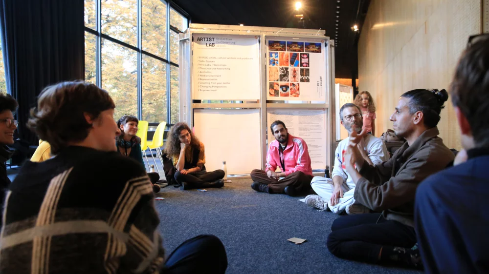 A group of people sits between exhibition walls with posters and graphics and discuss with each other.