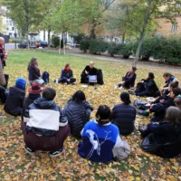 On a lawn covered with autumn leaves, about 20 people sit in a circle and listen to a speaker who holds an open tablet in one hand. Three people stand listening at the edge.