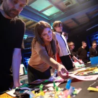 A group of people stand lined up around a large work table with documents and colored pieces of paper at the zine workshop. One of them reaches for a sheet.