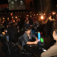 Two people in conversation at tables on the stage of the festival. In the background, a screen shows the faces of two participants in the conversation.