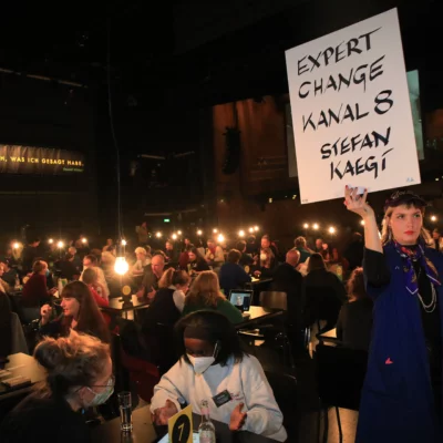 People are seated in pairs at around 50 symmetrically arranged tables in the stage area of the Berlin Festpsiele. A guest attendant holds up a large sign that reads "Expert Change: Kanal 8, Stefan Kaegi".