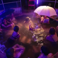 In einem beheizten Outdoor-Pool sitzen Menschen und sprechen miteinander. Eine Person hält einen aufgespannten Regenschirm in der Hand. Es ist am Abend und dunkel. In der Luft hängt der Dampf des warmen Wassers.