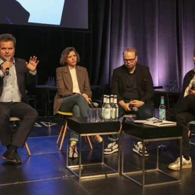 Vier Menschen sitzen auf einer Bühne in einer Podiumsdiskusion. Rechts ein Mann im Anzug, der in ein Mikrofon spricht. Links neben ihm eine Frau, ein Mann und eine weitere Frau, die konzentriert zuhören.