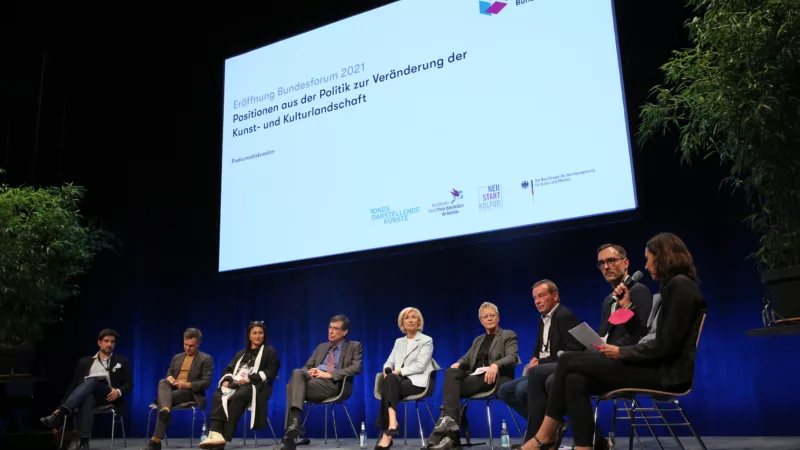 Neun Personen sitzen im Stuhlkreis zum Pdoiumsgespräch nebeneinander auf Stühlen auf dem Podium. Über ihnen ist eine große Leinwand zu sehen, auf der der Titel "Eröffnung Bundesforum 2021: Positionen aus der Politik zur Veränderung der Kunst- und Kulturlandschaft" zu lesen.