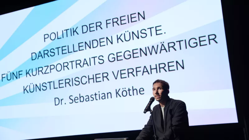 Serbastian Köthe hält an einem Redner*innenpult seinen Vortrag. Im Hintergrund ist auf einer Leinwand der Titel "Politik der Freien Darstellenden Künste. Fünf Kurzportraits gegenwärtiger künstlerischer Verfahren" zu lesen.