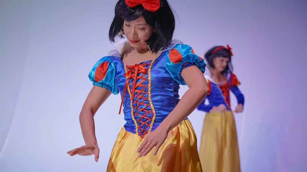 Im Vordergrund eine asiatisch gelesene Performerin im Schneewittchenkostüm. Im Hintergrund ist ein Double von ihr zu erkennen.