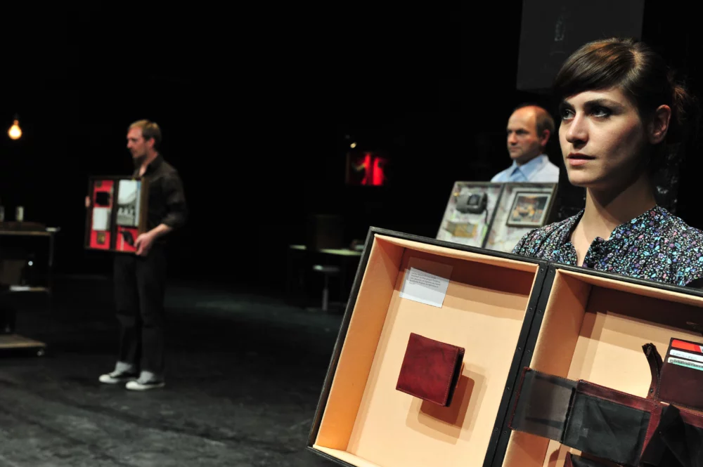 From a performance by Hofmann & Lindholm: three performers stand slightly offset on stage and hold a hinged box with showpieces in front of their chests.