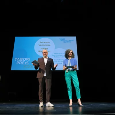 On stage, Annemie Vanackere and Holger Bergmann welcome the guests and open the evening. The moderator can be seen at the side of the picture.