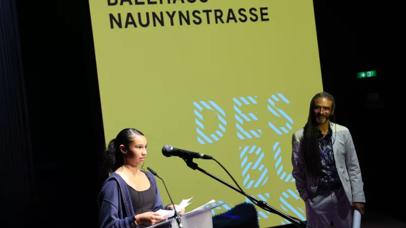 In the foreground a lectern. A teenager stands at it and gives a speech. The artistic director Wagner Carvalho stands in the background.