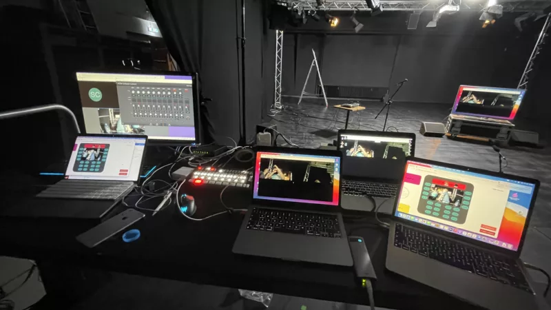 Blick vom Technikpult auf eine Blackbox-Bühne. Im Vordergrund stehen 4 Laptops und ein stationärer Bildschirm. Alle zeigen verschiedene PC-Programme, mit denen Einstellungen an einem Video vorgenommen werden.