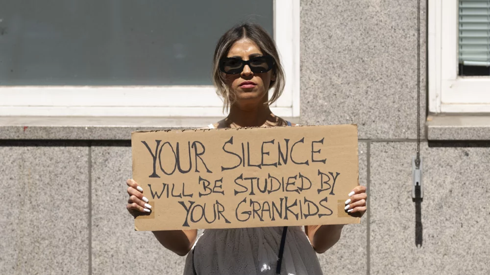 Eine Frau mit Sonnebrille hält vor ihrer Brust ein Pappschild mit den Worten "Your Silence will be studied by your Grankids", zu deutsch: "Deine/Eure Stille wird von Euren Enkelkindern studiert werden."