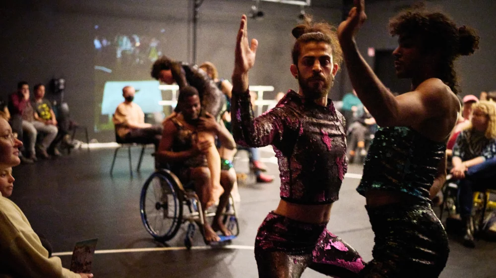 Two dancers in tight, colorful costumes stand opposite each other. Their knees are bent, their arms stretched upwards. In the background, a dancer in a wheelchair lifts a dancer. The audience sits on chairs at the edge of the scene.