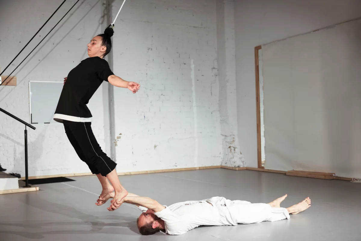 A man dressed in white is lying on his back on the floor of a studio. With outstretched arms, he reaches for the feet of a woman dressed in black, hanging from the ceiling by her hair. Her body is arched over, her arms outstretched.