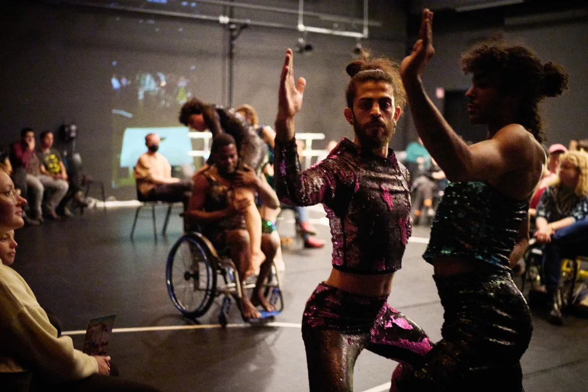 Zwei Tänzerinnen in engen, bunten Kostümen stehen sich gegenüber. Ihre Knie sind angewinkelt, die Arme nach oben gestreckt. Im Hintergrund hebt ein Tänzer im Rollstuhl eine Tänzerin hoch. Das Publikum sitzt auf Stühlen am Rande der Szene.