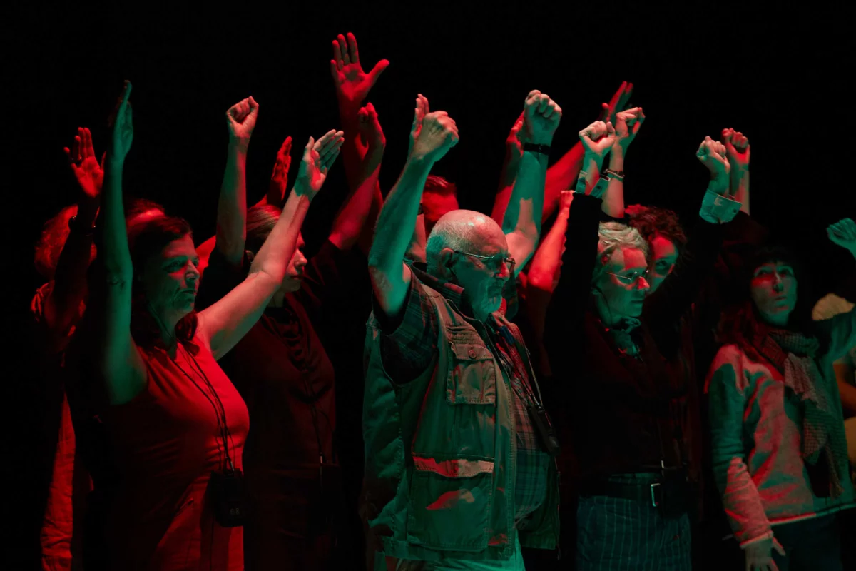 Bathed in subdued, reddish light, a group of people stand tightly packed in the room with their arms stretched upwards.