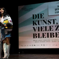 Vor der Plakatwand mit dem Veranstaltungstitel "DIE KUNST, VIELE ZU BLEIBEN" am Redner*innenpult stehend, hält die Autorin Şeyda Kurt ihren Impulsvortrag.