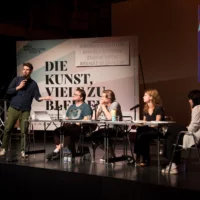 Vor einer Plakatwand mit dem Titel der Veranstaltung "DIE KUNST, VIELE ZU BLEIBEN" sitzen vier Menschen an einem Tisch auf der Bühne. Daneben stehend kündigt der Regisseur András Dömötör den Workshop an.