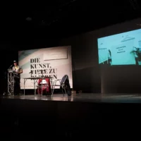 Jan Sowa steht für seinen Vortrag an einem Redner*innenpult vor einer Plakatwand mit dem Titel der Veranstaltungsreihe "DIE KUNST, VIELE ZU BLEIBEN". Im Hintergrund ist eine großflächige Videoprojektion zu sehen, die einen Grabstien mit der Aufschrift "Communism is dead" zeigt.