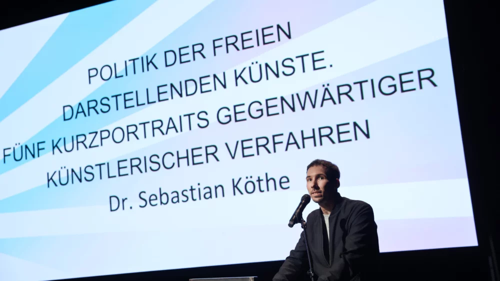 Serbastian Köthe gives his lecture at a lectern. In the background, the title “Politics of the Independent Performing Arts. Five short portraits of contemporary artistic processes".