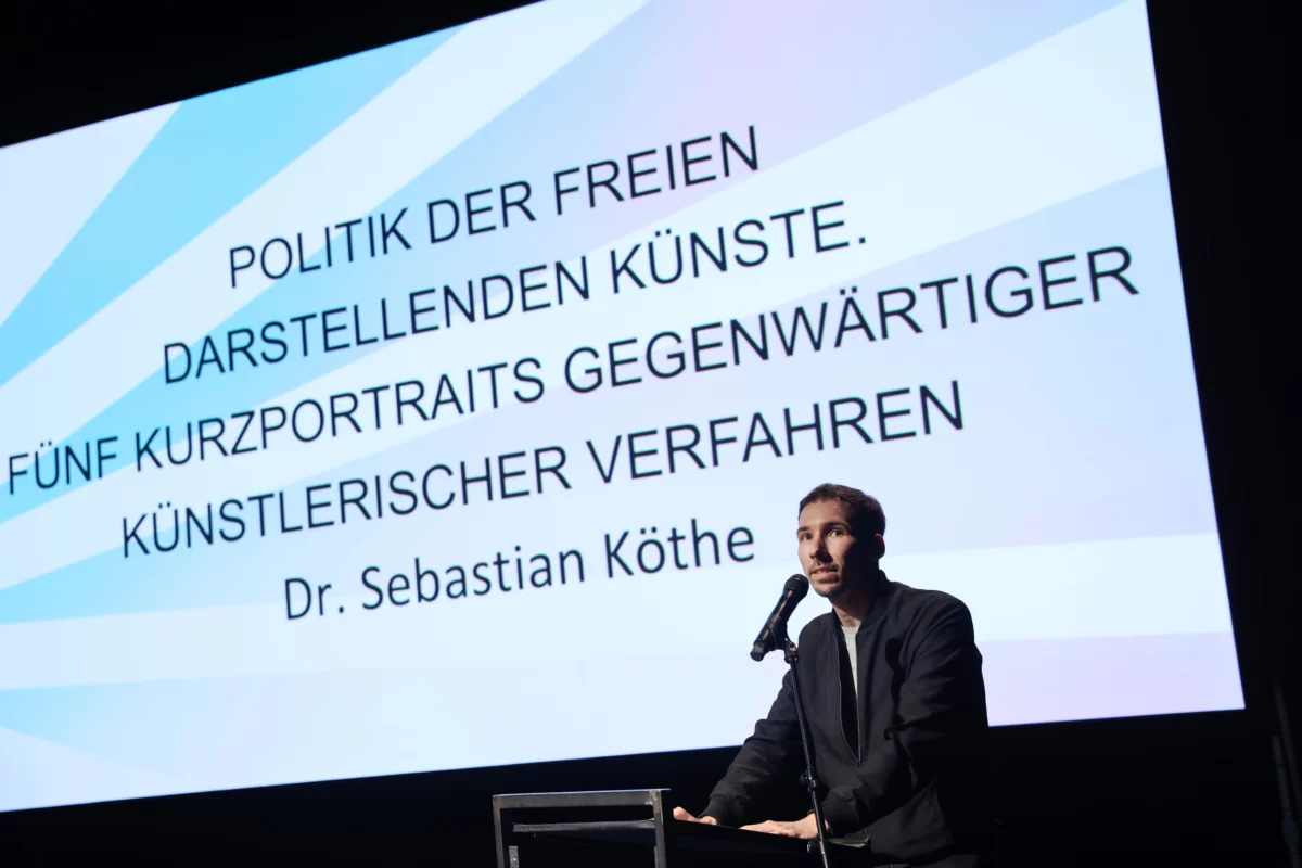 Serbastian Köthe hält an einem Redner*innenpult seinen Vortrag. Im Hintergrund ist auf einer Leinwand der Titel "Politik der Freien Darstellenden Künste. Fünf Kurzportraits gegenwärtiger künstlerischer Verfahren" zu lesen.
