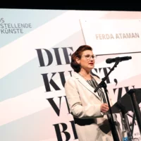 Ferda Ataman am Redner*innenpult dahinter der Aufsteller mit dem Logo von "DIE KUNST, VIEL ZU BLEIBEN".