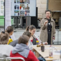 The artist and activist Tanja Krone stands in front of a group of workshop participants gathered at a table and gives a lecture with a microphone in her hand. The open truck stage with a monitor on it can be seen in the background.