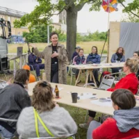 On an open meadow, surrounded by people sitting at long tables, the artist and activist Tanja Krone gives her workshop. In the background you can see an open truck, as well as a yellow brick building and trees behind it.