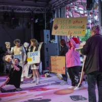 Seven performers of the play "Paul and Paula in search of happiness without end" stand in a semi-circle on stage, with the packed rows of spectators in the background. They carry various cardboard signs in front of them that read "Food without restrictions, cake for all", "In love forever and ever, for justice, in peace" and "Checkout 1 - BILLA".