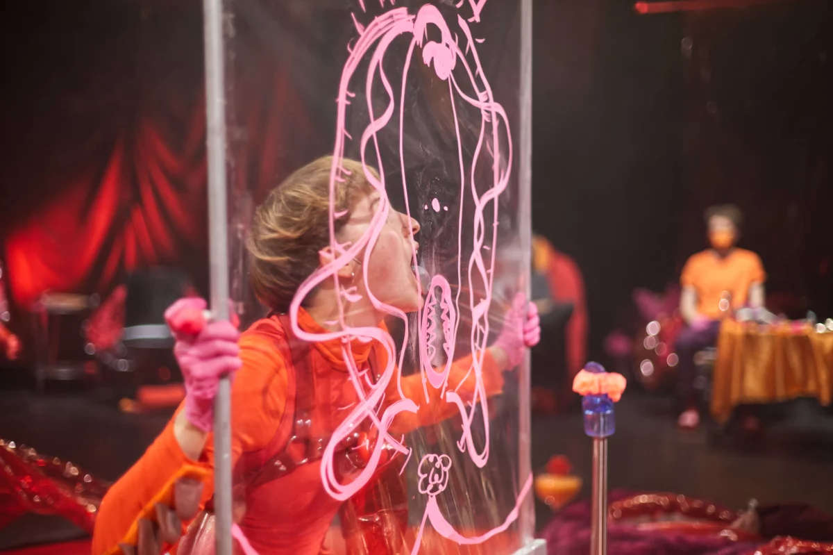 Eine in auffälligem orange gekleidete Person hält mit beiden Händen eine Plexiglasscheibe vor sich, auf der mit pink eine Vagina aufgezeichnet ist, und leckt mit ihrer Zunge die Scheibe ab.