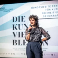 Felizitas Stilleke steht vor einer Leinwand mit der Aufschrift "DIE KUNST, VIELE ZU BLEIBEN". Sie spricht in ein Mikrofon, das sie in der Hand hält.