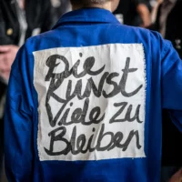 Rear view of a person in overalls. A fabric with the inscription "DIE KUNST, VIELE ZU BLEIBEN" is sewn onto the overalls.