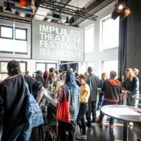 Many people are standing in the foyer of the FFT, engrossed in conversation. The projection on a screen: Impulse Theaterfestival 29.5.-9.6.2024