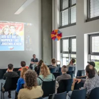 Arne Vogelgesang sitzt vor einem Publikum, hinter ihm eine Projektion mit einem Poster, auf dem steht: Heimat & Identität gibt's nur bei uns: AfD ist die Nummer 1 bei Jugendlichen." Darunter das Logo der AfD.