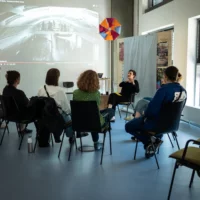 Anna Lux speaks to the workshop participants sitting in a semi-circle in front of her.Behind her is a projection of a music video.