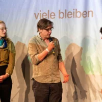 Standing side by side on the stage, Anne-Cathrin Lessel, Thomas Frank and Felizitas Stilleke open the Forum in Leipzig with a joint greeting. A section of a video projection with the words "remain many " can be seen in the background.