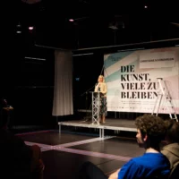 Dr. Christiane Schenderlein, member of the Bundestag and cultural policy spokesperson for the CDU, stands in front of a billboard with the inscription "DIE KUNST, VIELE ZU BLEIBEN" and gives a keynote speech to the audience while standing at a lectern.