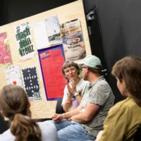 People sit in a semi-circle in front of a poster wall with various poster motifs and discuss with each other.